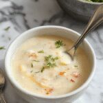 A creamy, pale beige cauliflower soup served in a rustic white bowl, garnished with fresh chopped herbs and a drizzle of olive oil, with a side of toasted gluten-free bread slices on a wooden table.