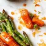 A vibrant plate featuring honey-glazed carrots and green beans, beautifully arranged with a glossy finish. The carrots are sliced into rounds, coated in a shiny honey glaze, and garnished with a sprinkle of fresh herbs. The green beans are tender, bright green, and slightly caramelized, complementing the carrots with their fresh, crisp appearance. The dish is presented on a rustic white plate, set against a wooden table with a soft, natural lighting highlighting the glossy textures and colorful presentation.
