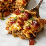 A bubbling, golden-brown casserole topped with crispy bacon and melted cheese, garnished with fresh chopped herbs. The dish is served in a rustic white ceramic baking dish, revealing layers of seasoned ground beef, cheese, and bacon, with a fork lifting a portion to showcase its cheesy, savory interior.