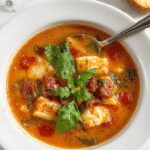 A steaming bowl of Mediterranean soup with vibrant colors, featuring chopped tomatoes, zucchini, chickpeas, and fresh herbs, garnished with a drizzle of olive oil and a sprig of basil, served in a rustic white bowl on a wooden table.