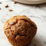 A close-up of warm pumpkin muffins topped with chocolate chips, arranged on a rustic wooden platter. The muffins have a golden-brown crust with melted chocolate chips visible on top, accompanied by a scattering of pumpkin seeds and a hint of cinnamon in the background.