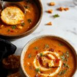 A steaming bowl of Witches Soup with rich, dark broth topped with vibrant herbs and garnishes, surrounded by mystical ingredients on a rustic wooden table, with a glowing candle and enchanted symbols in the background, enhancing the cozy and witchy atmosphere.