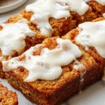 A slice of pumpkin bread topped with a generous swirl of creamy white cream cheese frosting, presented on a rustic wooden plate. The bread has a warm, golden-brown crust with visible specks of spice and pumpkin flecks. The frosting appears thick, smooth, and luscious, contrasting beautifully with the moist, dense interior of the bread in the cross-section. In the background, a whole pumpkin and a drizzle of frosting are softly blurred, enhancing the cozy, autumnal appeal of the scene.