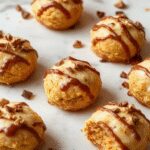 A close-up of several pumpkin-shaped cheesecake truffles coated in orange and white striped candy melts, presented on a rustic wooden platter with cinnamon sticks and small decorative pumpkins in the background. The truffles have a smooth, glossy surface with visible specks of graham cracker crumb texture underneath the vibrant orange coating.