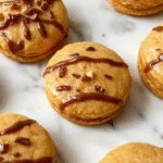 A tray of vibrant orange pumpkin pie macarons with smooth, glossy shells and ruffled edges, neatly arranged on a rustic wooden surface with scattered cinnamon and pumpkin seeds in the background.