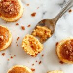 A close-up shot of the Pumpkin Spice Cheesecake Bites arranged on a white platter. The bites are creamy, with a hint of orange pumpkin and topped with a sprinkle of cinnamon or nutmeg. The presentation highlights their smooth texture and inviting, bite-sized portion sizes, accented by a few decorative fall leaves in the background.