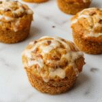 A close-up of a freshly baked pumpkin swirl muffin, showcasing its moist, golden-brown crust with a visible swirl of creamy pumpkin filling. The muffin rests on a rustic wooden surface, garnished with a light dusting of cinnamon and small pumpkin seeds around it. The vivid orange and warm tones highlight the inviting, autumnal appeal of the dessert.