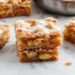 A close-up of golden-brown apple pie bars sliced to reveal a juicy, cinnamon-spiced apple filling nestled inside flaky pastry crust. Garnished with a dusting of powdered sugar and a sprig of fresh mint, the bars are presented on a rustic wooden platter against a cozy fall-themed background with hints of cinnamon sticks and whole apples.