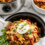 A cozy, bubbling taco casserole in a rustic ceramic slow cooker, topped with melted cheese, fresh chopped cilantro, and diced tomatoes. The dish showcases a rich, hearty mixture of seasoned ground beef, beans, and corn, with steam rising and a vibrant, colorful presentation highlighting the melting cheese and garnishes.