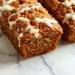 A freshly baked loaf of Snickerdoodle Pumpkin Bread sits on a rustic wooden table, topped with a cinnamon-sugar coating. The bread's golden crust is slightly cracked, revealing a moist, pumpkin-rich interior. A scattering of cinnamon sugar and cinnamon sticks surround the loaf, adding warmth to the scene. Natural lighting emphasizes the textures and inviting aroma.