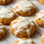 A close-up of soft pumpkin cookies topped with smooth cream cheese icing, arranged on a rustic ceramic plate. The cookies are golden brown, with a slightly cracked surface, and the icing is generously spread, creating a luscious, inviting look. The background features a cozy autumn setting with hints of fall leaves and a warm-toned table.