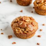 A close-up of freshly baked spiced pear muffins arranged on a rustic wooden platter. The muffins have a golden-brown crust with visible chunks of tender pear and specks of cinnamon. The muffins are garnished with a light dusting of powdered sugar and accented by a few slices of fresh pear and cinnamon sticks in the background. The scene is set on a cozy kitchen table with a warm, inviting ambiance.