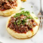 A rich, rustic bowl of short rib ragu featuring tender, shredded beef in a dark, savory sauce, garnished with fresh herbs, served over a creamy mound of silky mashed potatoes. The dish is presented on a white ceramic plate with a sprig of thyme, steamy and inviting, with a side of warm bread and a glass of red wine nearby.
