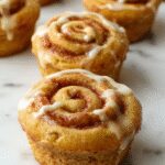 A close-up of golden-brown pumpkin cinnamon roll muffins arranged on a rustic wooden tray, with swirls of cinnamon and pumpkin peeking through the tops and a drizzle of icing on some muffins, set against a cozy fall-themed backdrop with autumn leaves.