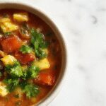 A steaming bowl of Tuscan Vegetable Soup garnished with fresh basil and a drizzle of olive oil. The soup contains colorful vegetables like tomatoes, zucchini, and spinach, with visible chunks of hearty beans and pasta, served on a rustic wooden table with crusty bread on the side.